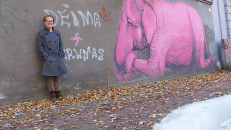 Laura vom Achtsam Reisen Festival lehnt an einer Mauer, auf die ein großer rosa Elefant gemalt wurde.