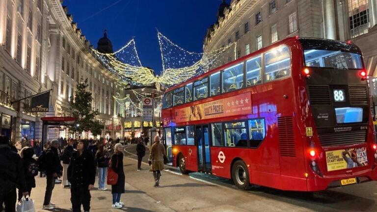 In den vielen Einkaufsstraßen in London hängen winterliche Lichter, zum Beispiel leuchtende Engel in der Regent Street. Ein roter Bus fährt vorbei.