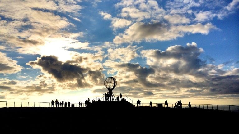 Am Nordkap geht die Sonne im Sommer niemals unter. Während die am blauen Himmel hell scheint sind die Menschen auf dem Bild ganz dunkel.